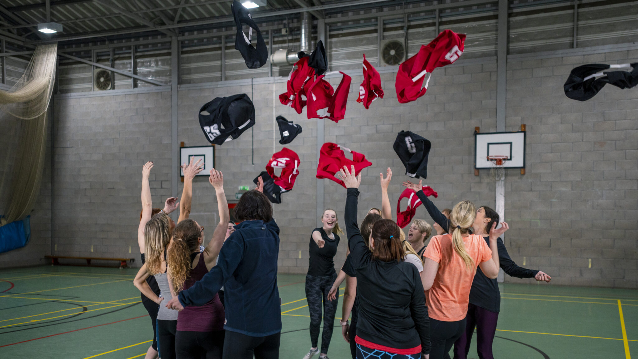 A wide shot of a group of women wearing sports clothing and netball bibs in a sports hall in Newcastle Upon Tyne. They are all huddled together and throwing their netball bibs up in the air.